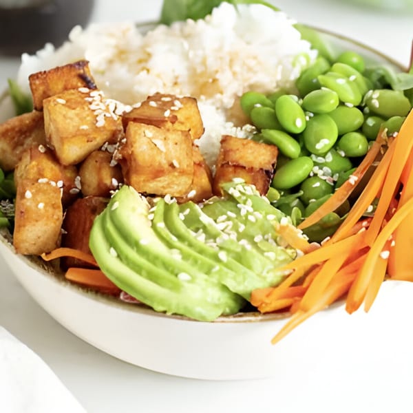 A vibrant poke bowl with crispy tofu, white rice, edamame, sliced avocado, and shredded carrots.